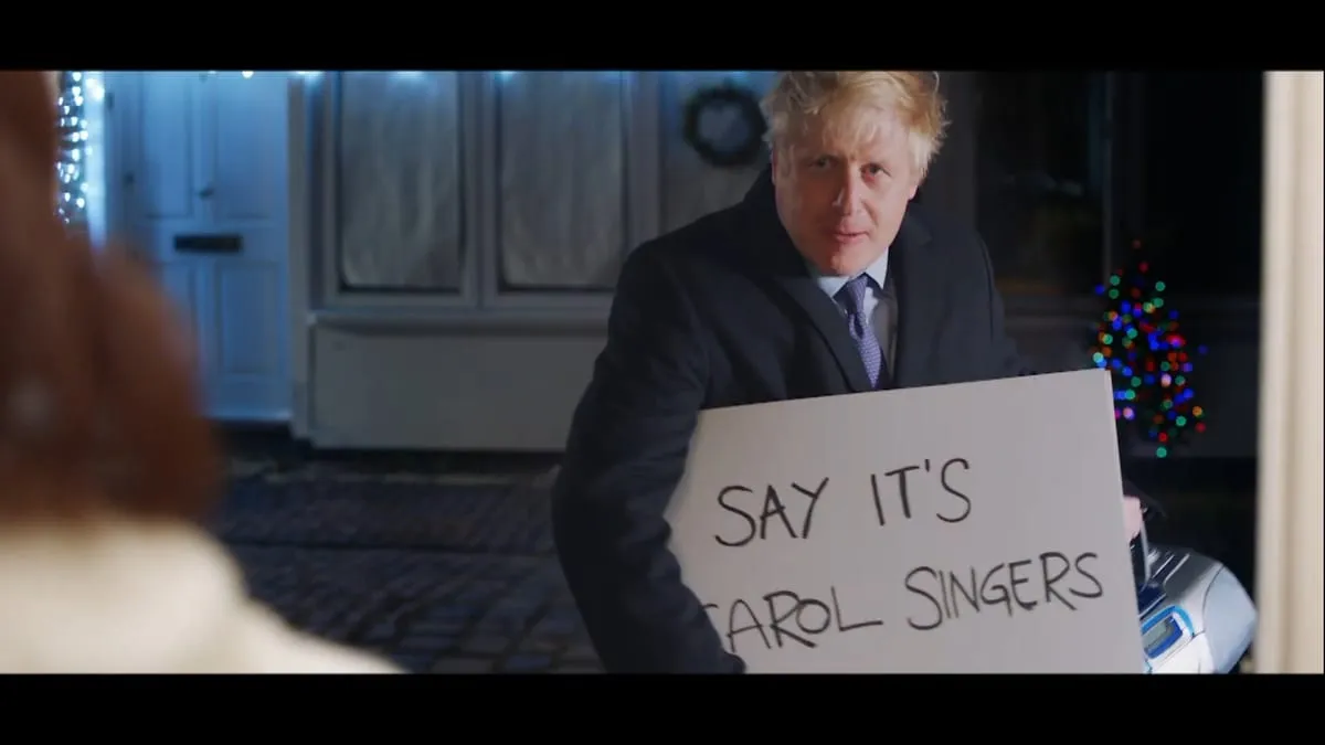 Still from Topham Guerin’s Love Actually parody ad: Boris Johnson holds a cue card reading “Say it’s carol singers”.