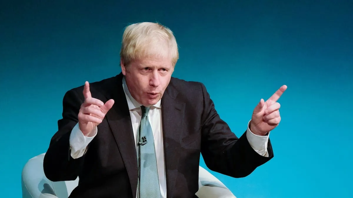 Boris Johnson gesturing during a speech against a teal backdrop.